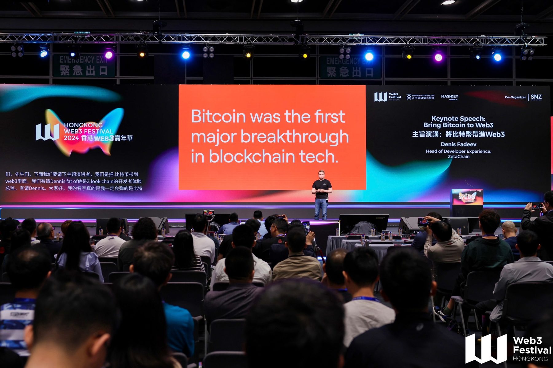 Denis on stage giving a keynote to a full hall, the slide reads “Bitcoin was the first major breakthrough in blockchain tech”